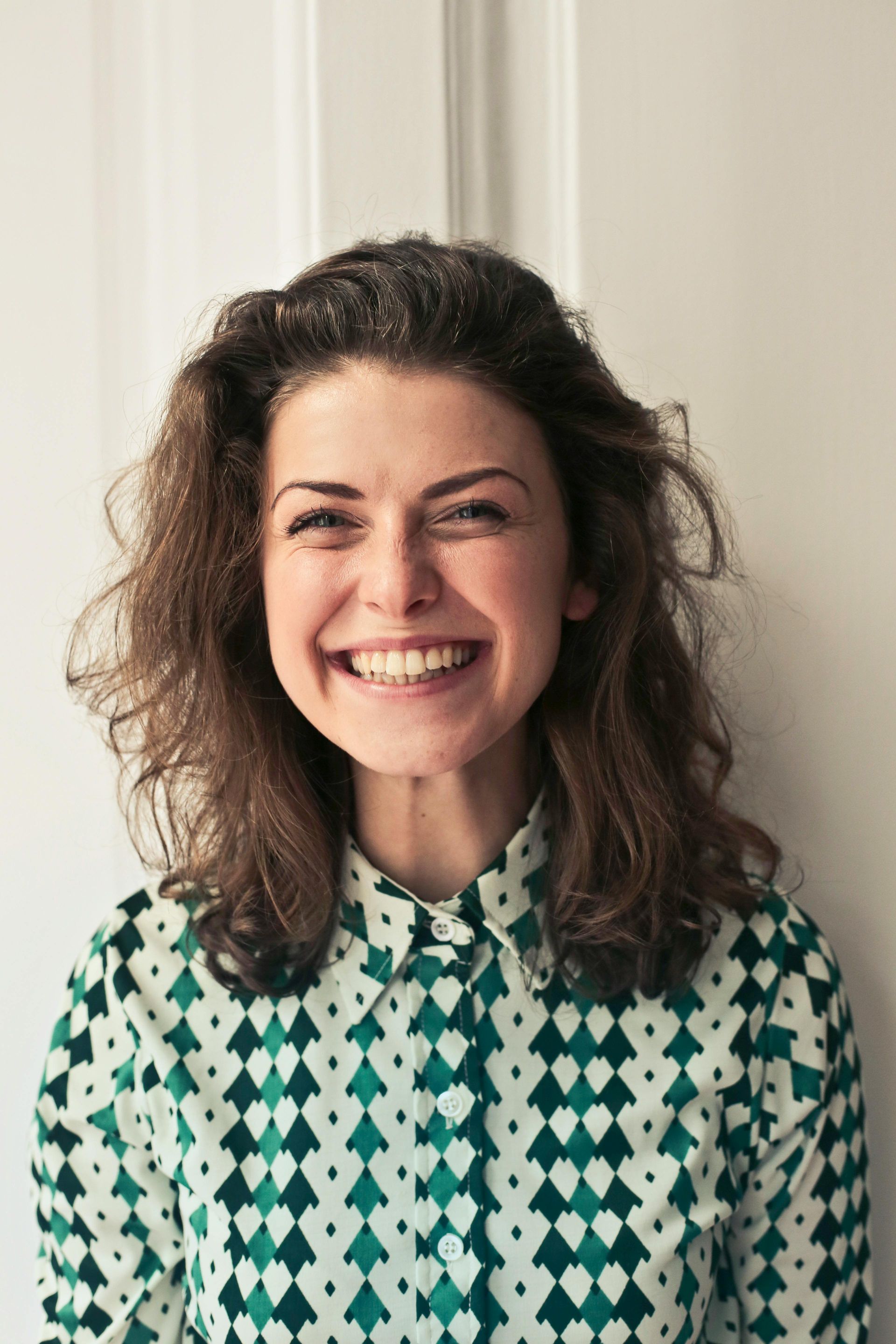 Woman with curly brown hair smiles broadly, wearing a green and white patterned shirt.